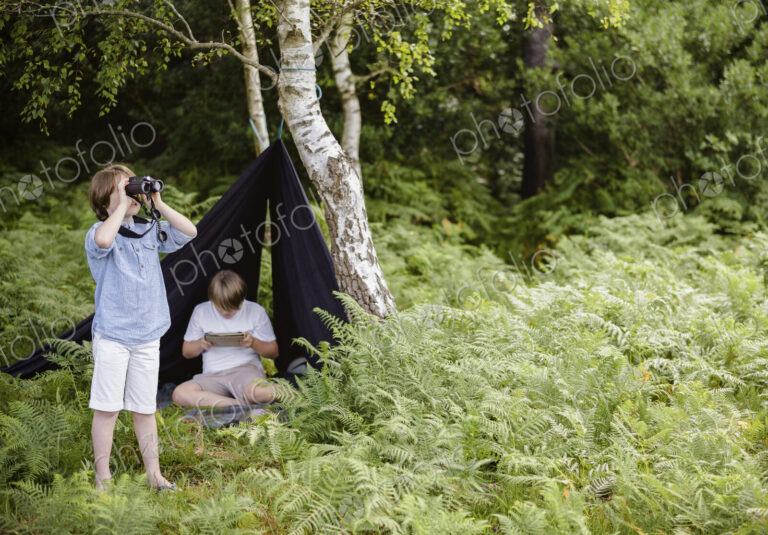 Two boys camping in New Forest. One sitting under a black canvas shelter. One boy…
