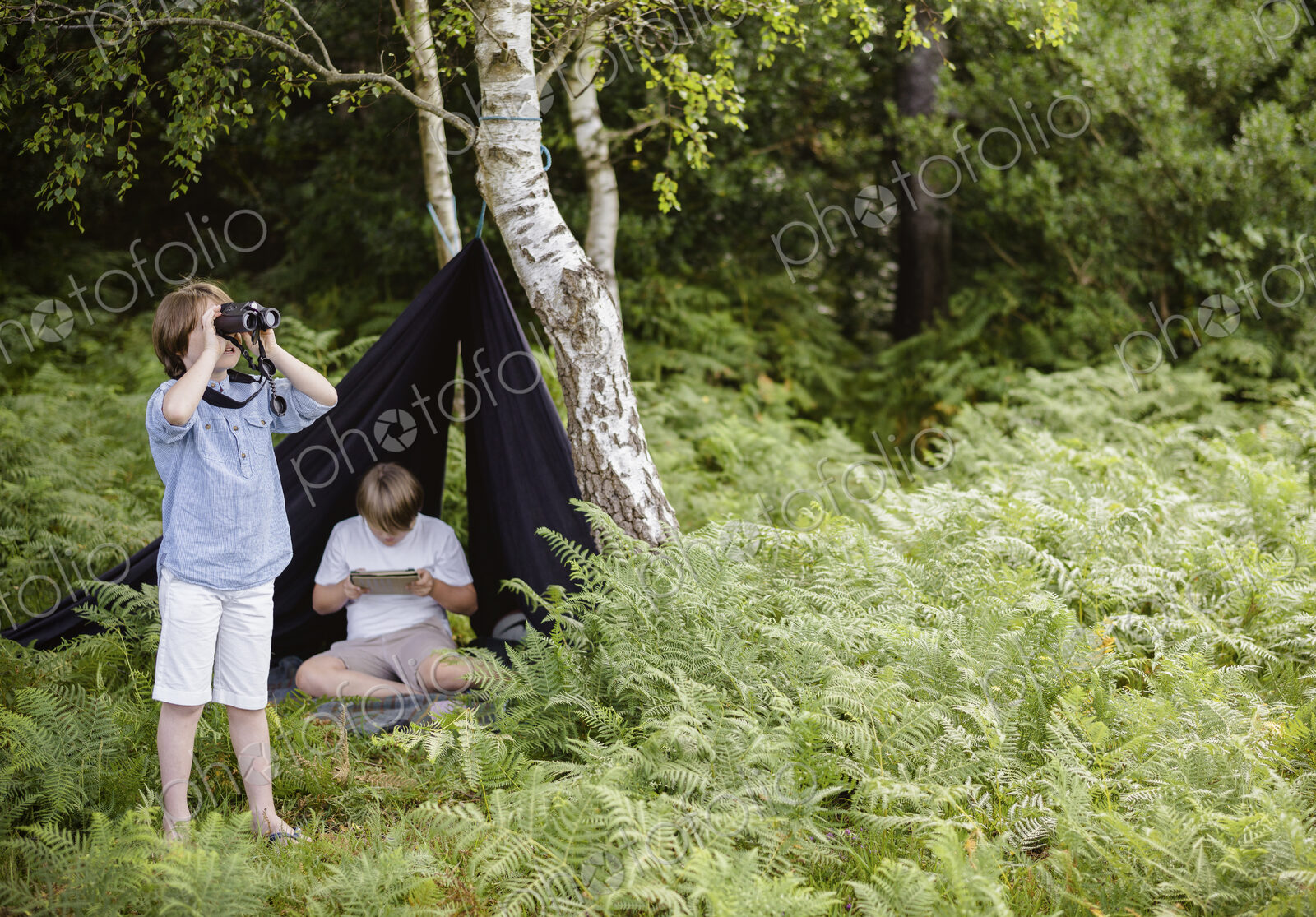 Two boys camping in New Forest. One sitting under a black canvas shelter. One boy…