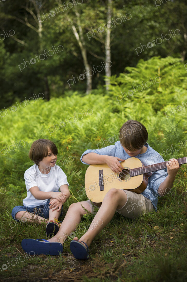 Two boys camping in New Forest. Sitting on the grass, one playing a guitar.