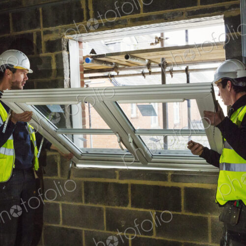 Two builders in hard hats and high visibility jacket fitting a window frame.