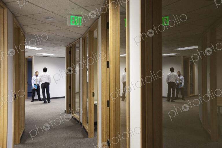 Two business men standing and talking at the end of an office hallway.