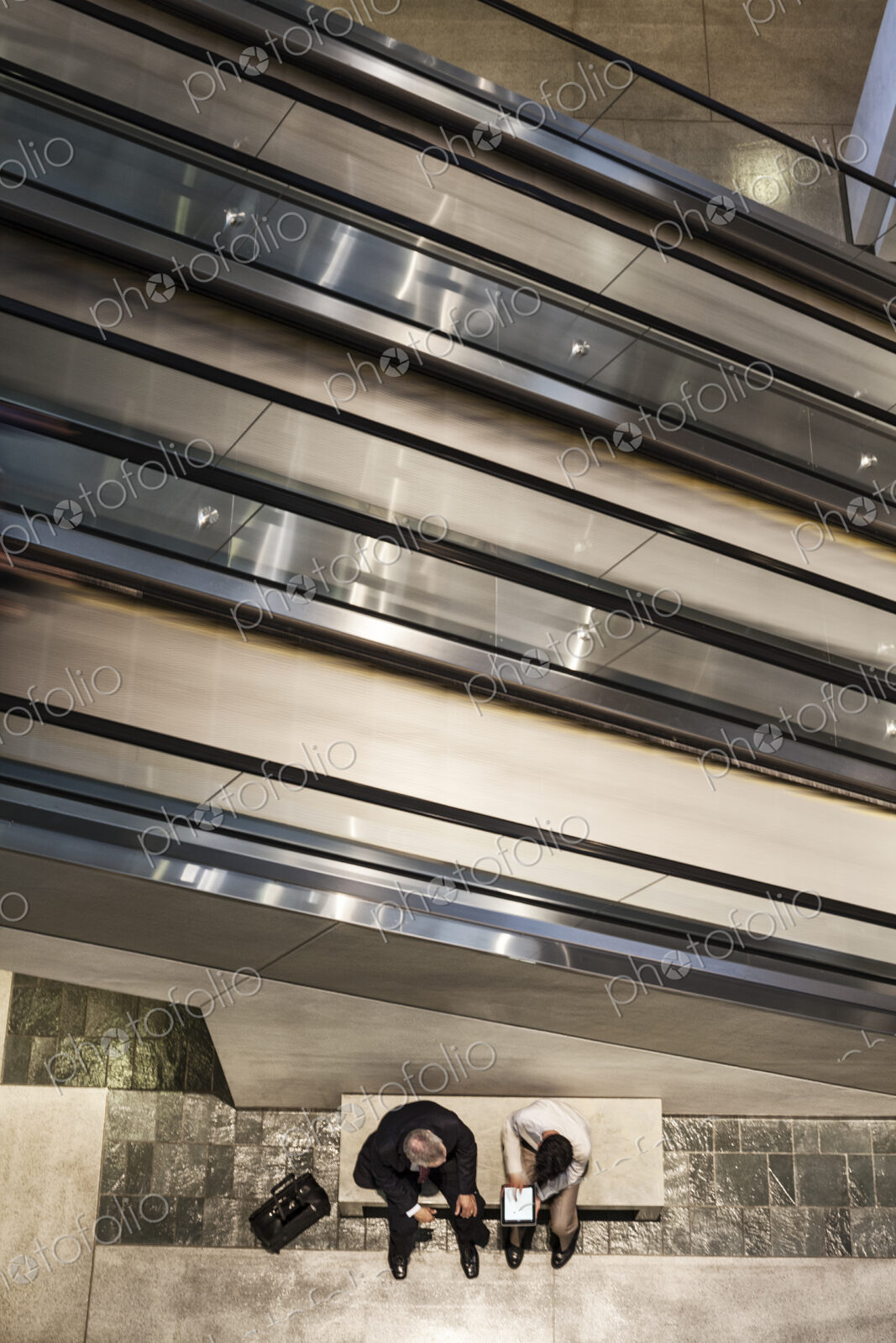 Two businessmen on a notebook computer near an escalator.