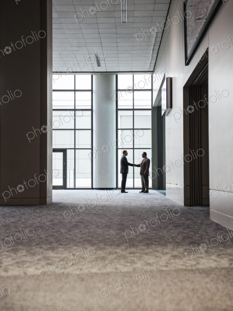 Two businessmen silhouetted and shaking hands in front of a large convention center…