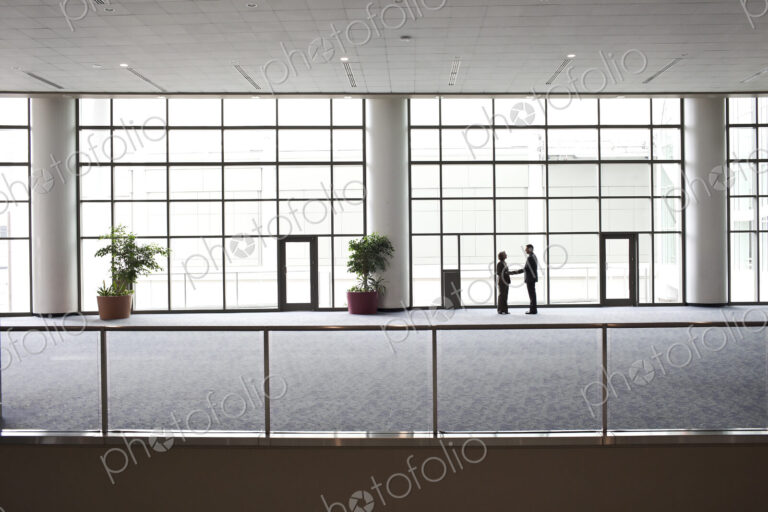 Two businessmen silhouetted and shaking hands in front of a large convention center…