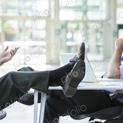 Two businessmen talking at a desk with one person's feet resting on top of the table Two businessmen talking at a desk with one person's feet resting on top of the table