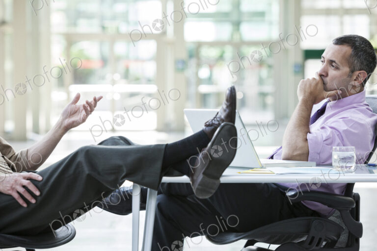 Two businessmen talking at a desk with one person's feet resting on top of the table
