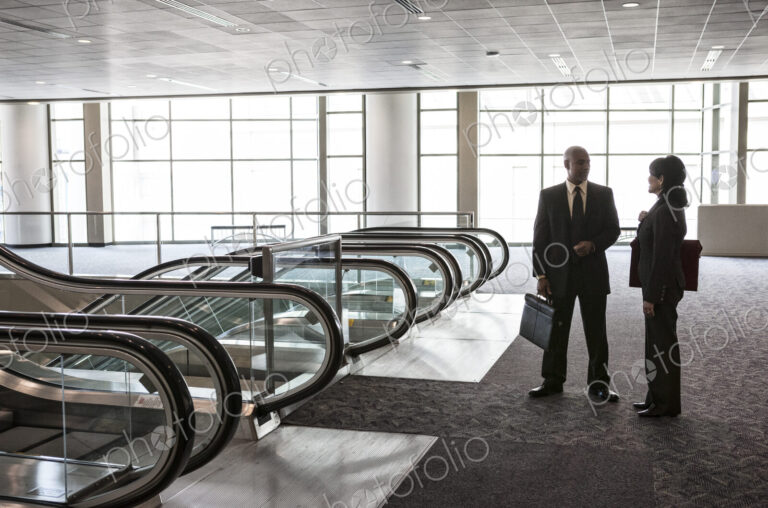 Two businesspeople, a man and woman standing talking by a bank of escalators in a…