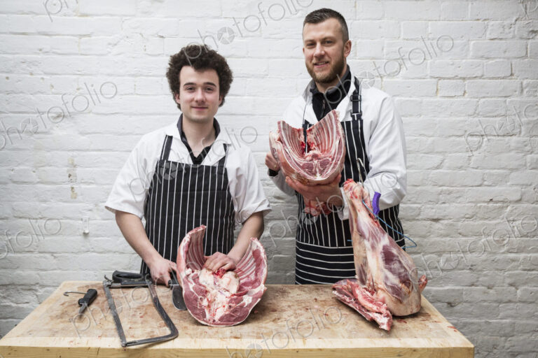 Two butchers side by side holding meat joints, part of a large butchered carcass.
