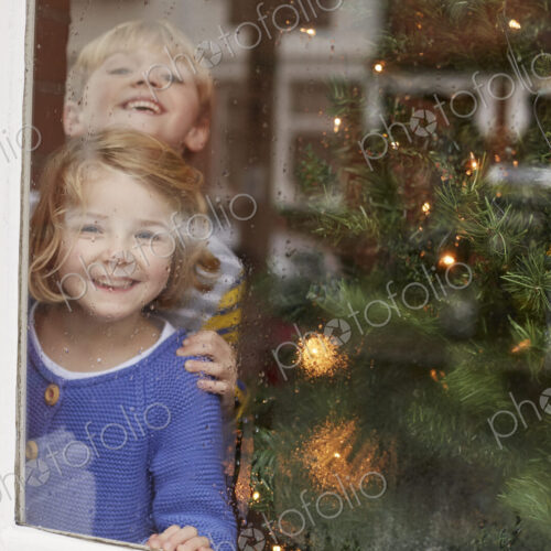 Two children, a boy and girl, looking out of a window at home beside a Christmas tree.