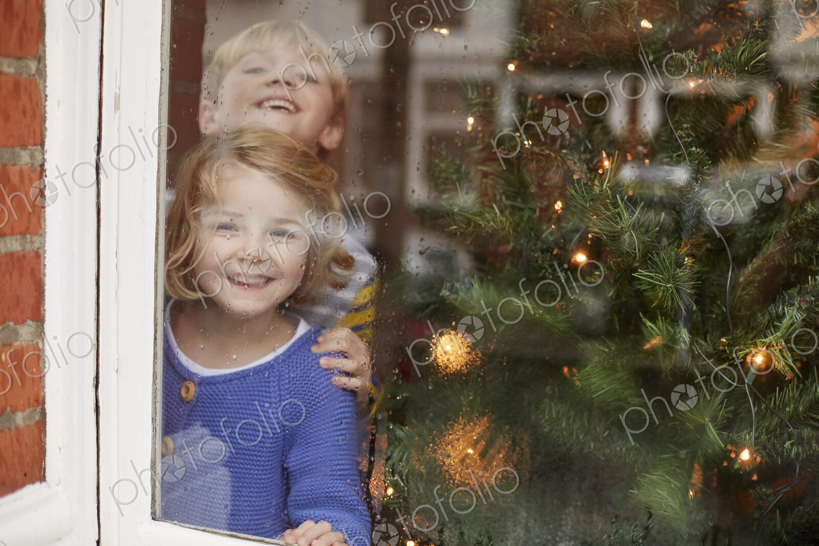 Two children, a boy and girl, looking out of a window at home beside a Christmas tree.
