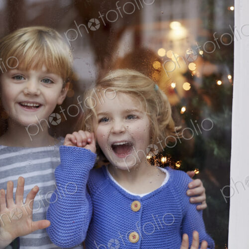 Two children, a boy and girl, looking out of a window at home with excited expressions.