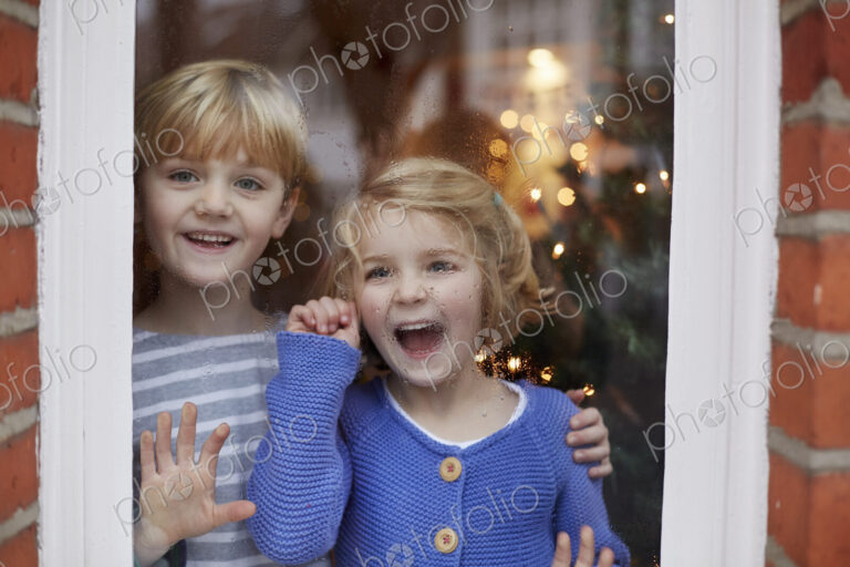 Two children, a boy and girl, looking out of a window at home with excited expressions.