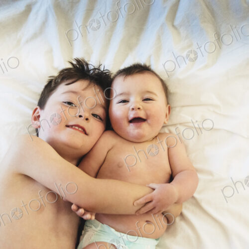 Two children, a brother and baby sister lying down playing together.