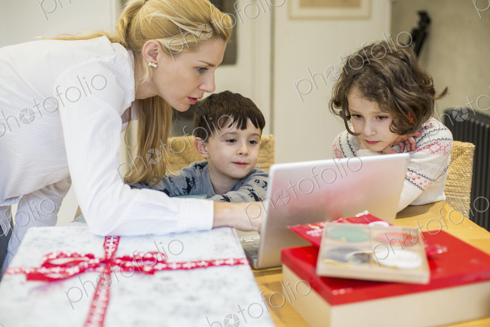 Two children and their mother looking at a laptop computer screen around a kitchen table.