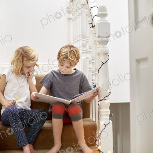 Two children seated side by side on the stairs at home, reading a book together. Two children seated side by side on the stairs at home, reading a book together.
