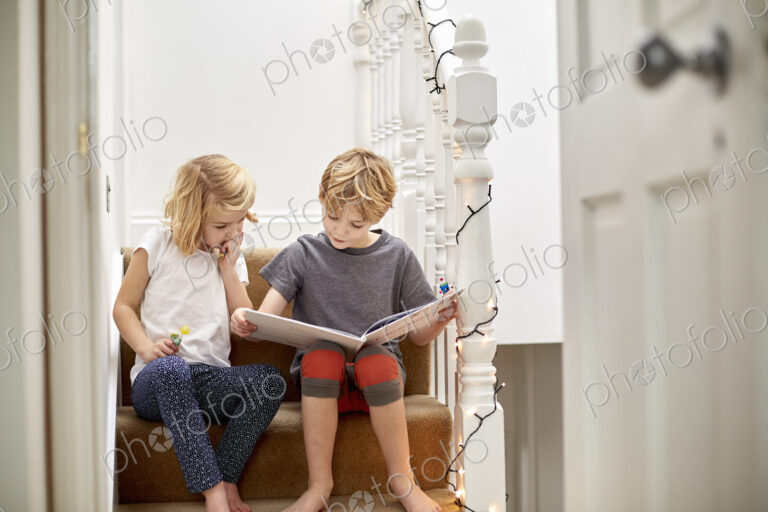 Two children seated side by side on the stairs at home, reading a book together.
