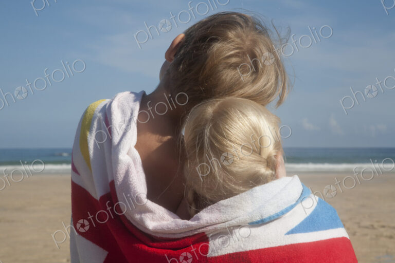 Two children sharing a towel. Back view of a brother and sister sitting on the beach…