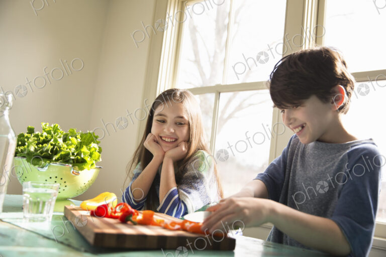 Two children sitting at a table. A boy using a knife and chopping bell peppers on a board.