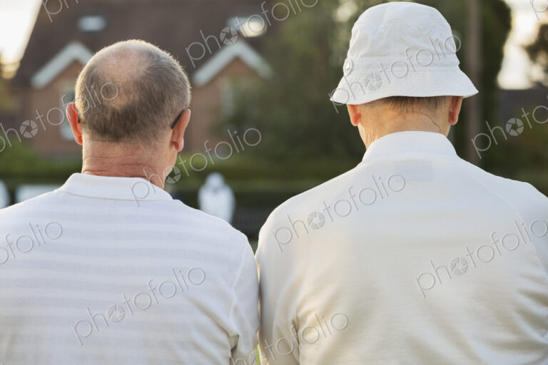 Two companions, two men standing side by side in white shirts outdoors. Lawn bowls…
