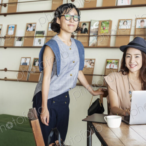 Two female Japanese professionals at a table in a co-working space, using laptop computer.