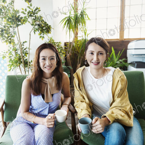 Two female Japanese professionals sitting in a co-working space, smiling at camera. Two female Japanese professionals sitting in a co-working space, smiling at camera.