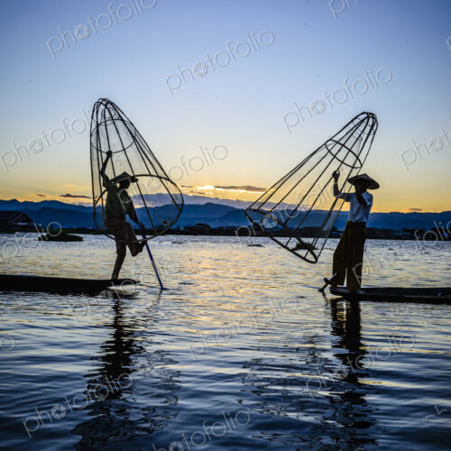 Two fishermen standing on their boats fishing on Lake Inle at dusk.
