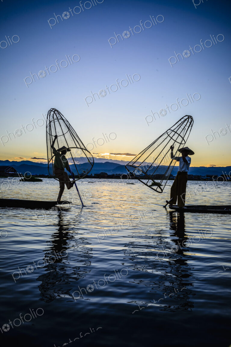Two fishermen standing on their boats fishing on Lake Inle at dusk.