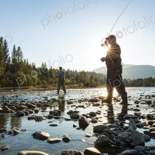 Two fly fisherman standing in rocky riverbed, casting their fishing rods. Two fly fisherman standing in rocky riverbed, casting their fishing rods.