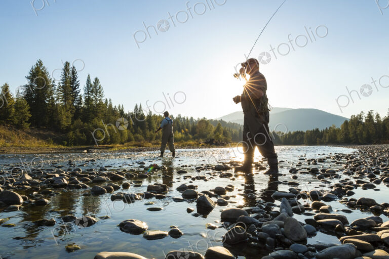Two fly fisherman standing in rocky riverbed, casting their fishing rods.