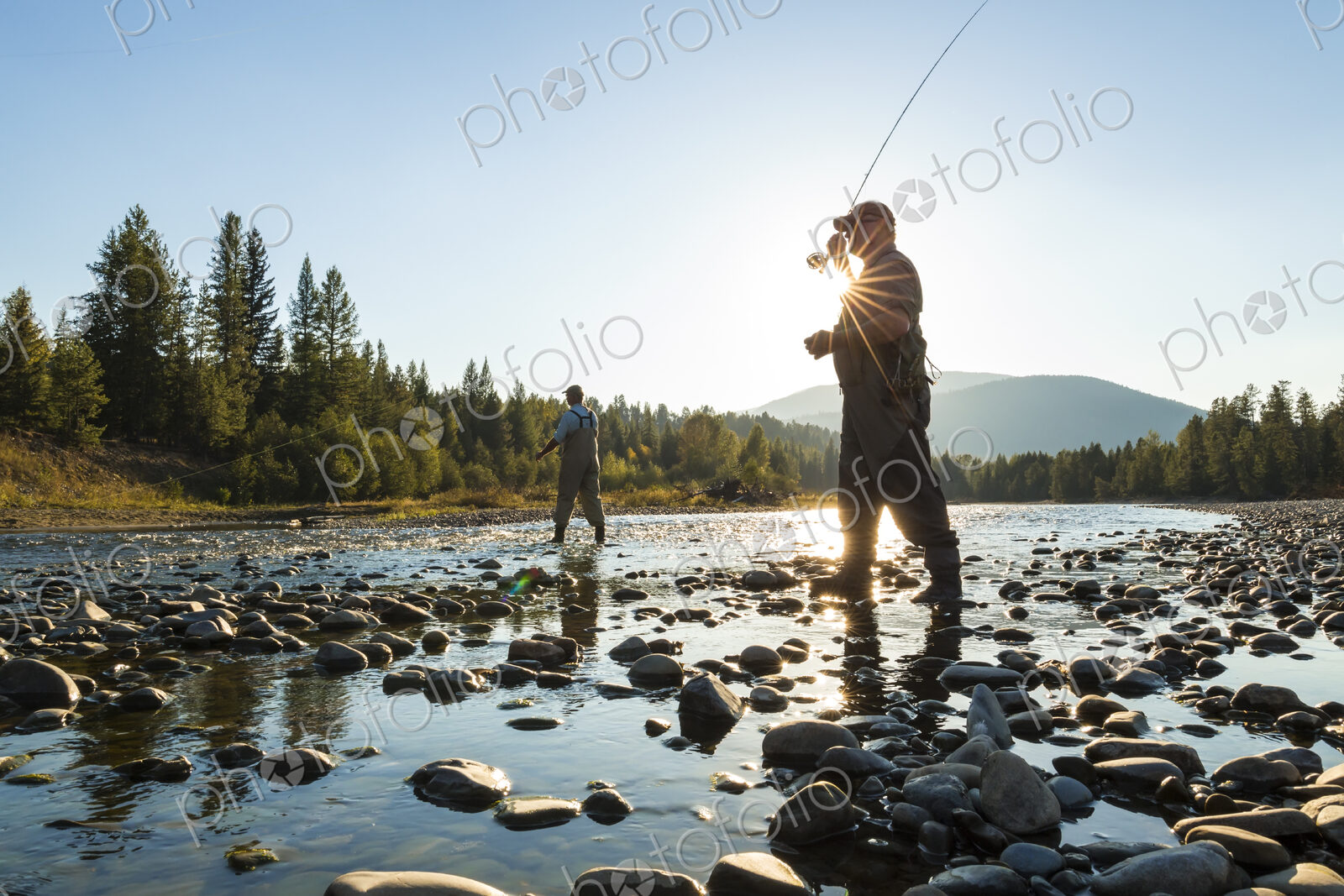 Two fly fisherman standing in rocky riverbed, casting their fishing rods.