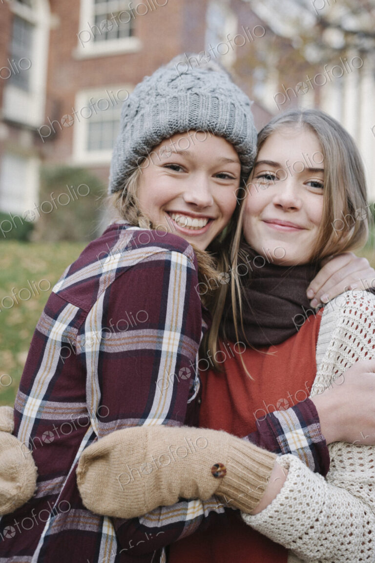 Two girls in hats and gloves wrapped up for cold weather, hugging and laughing.