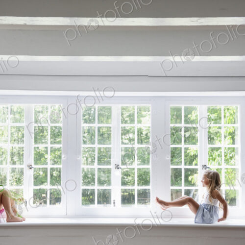 Two girls playing, sitting on a ledge by a large window.