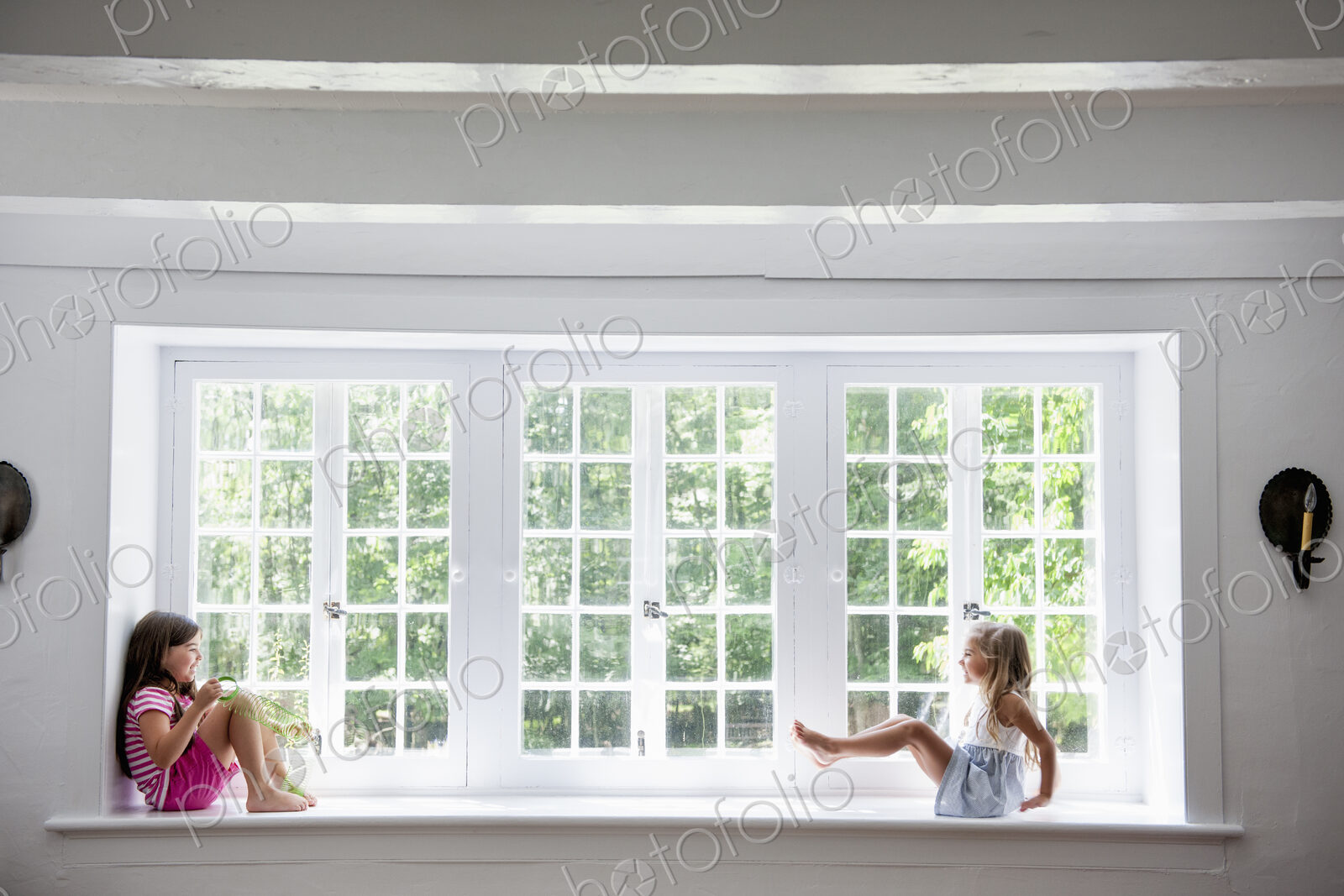 Two girls playing, sitting on a ledge by a large window.