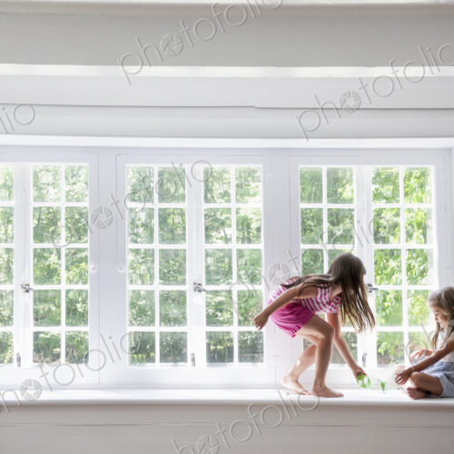 Two girls playing, sitting on a ledge by a large window.