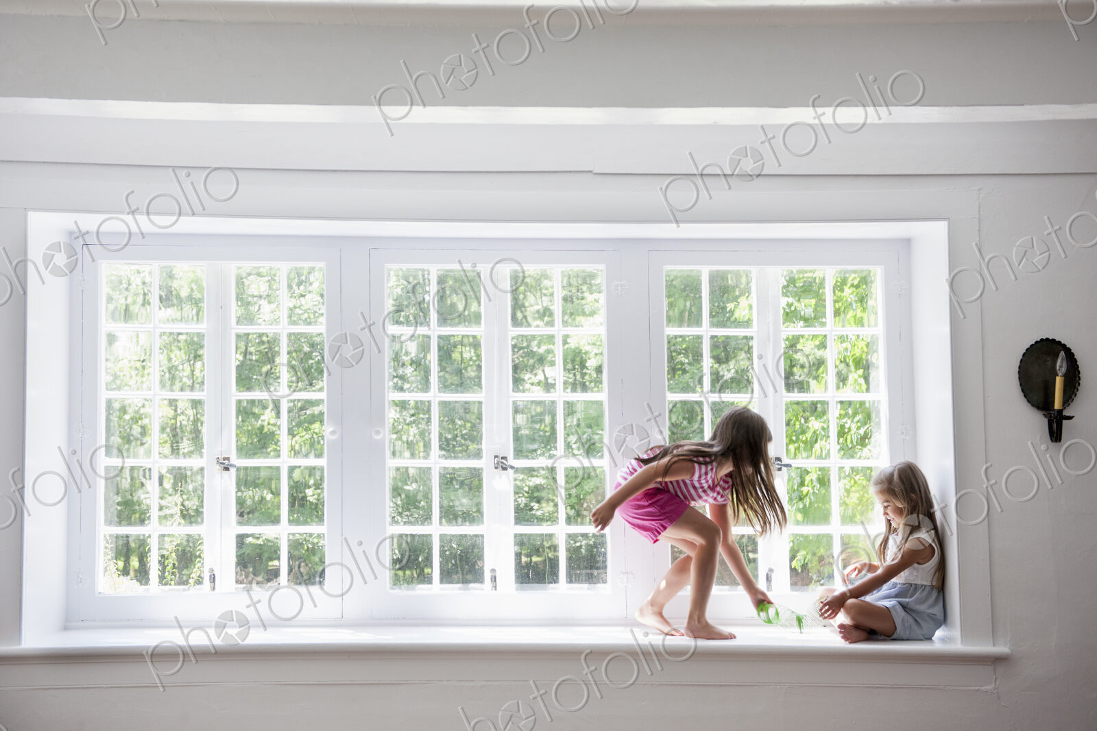 Two girls playing, sitting on a ledge by a large window.