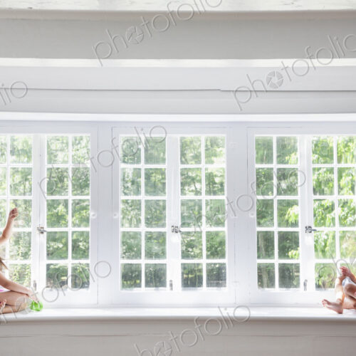 Two girls playing, sitting on a ledge by a large window.