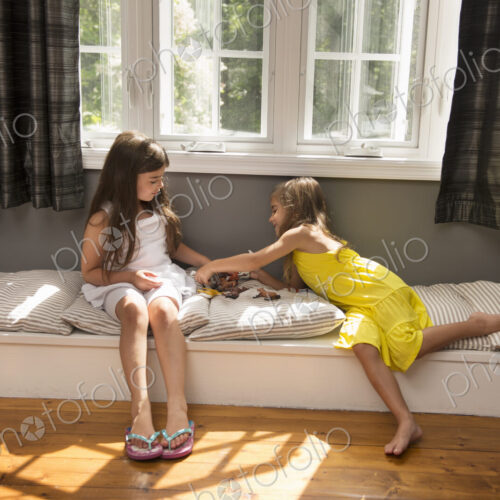 Two girls playing together, sitting on a window seat indoors.