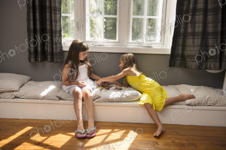 Two girls playing together, sitting on a window seat indoors.