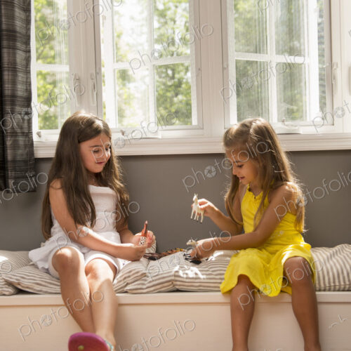 Two girls playing together, sitting on a window seat indoors.