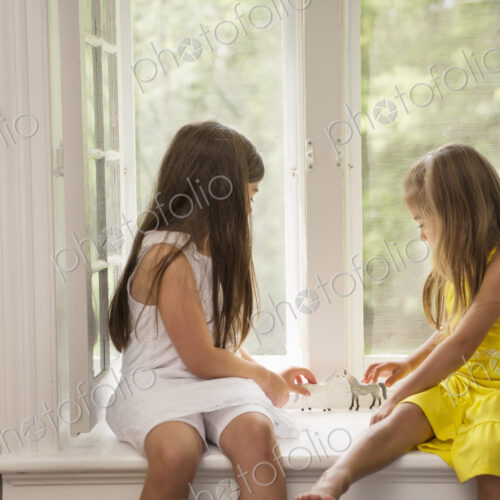 Two girls playing together, sitting on a window seat indoors.