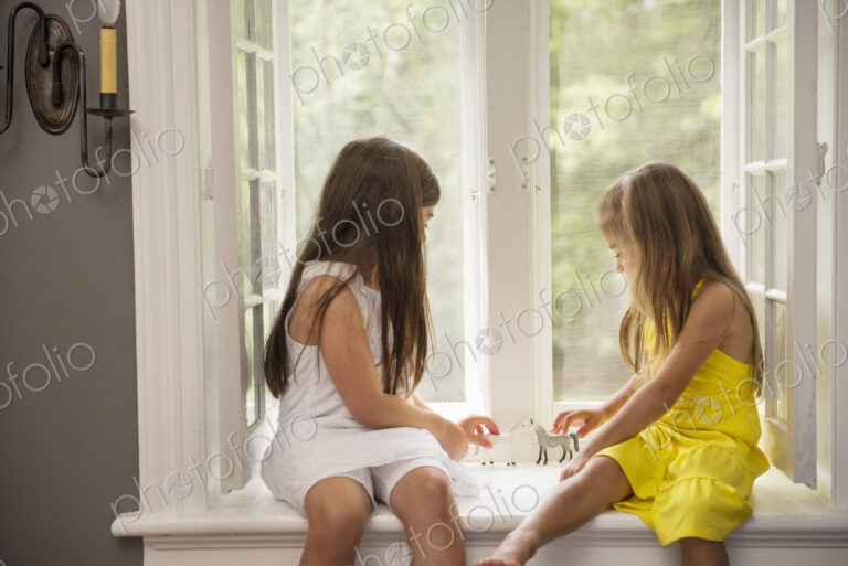 Two girls playing together, sitting on a window seat indoors.