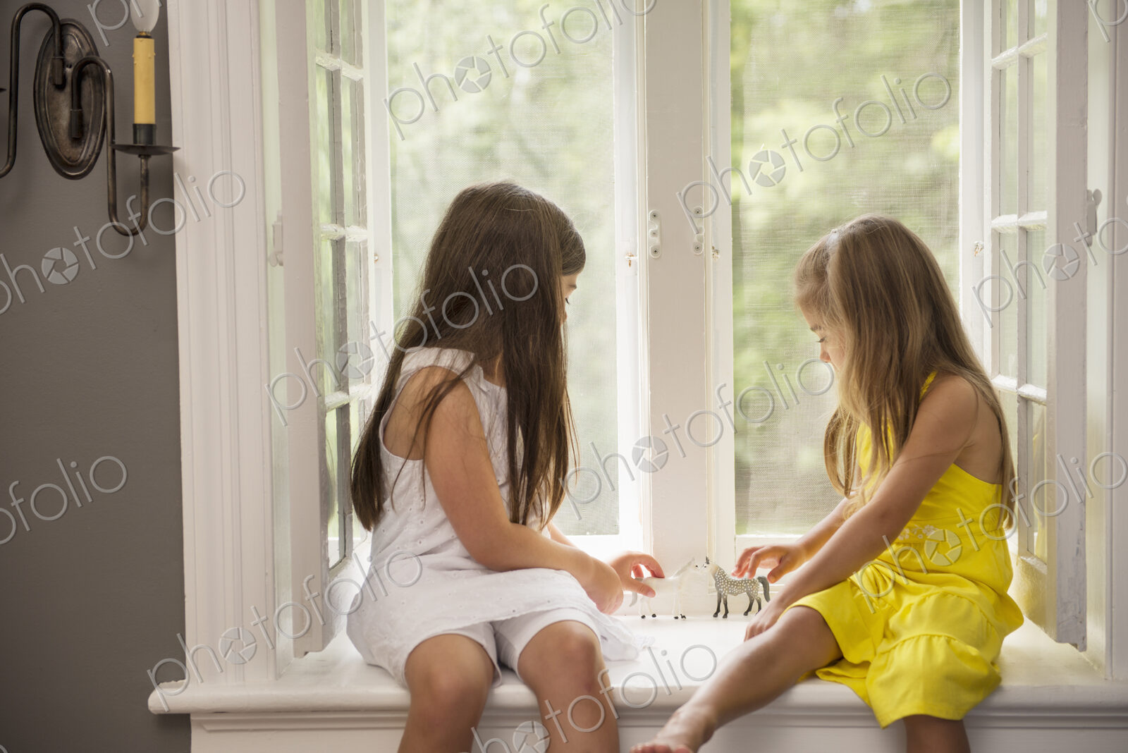 Two girls playing together, sitting on a window seat indoors.