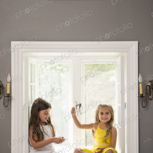 Two girls playing together, sitting on a window seat indoors.