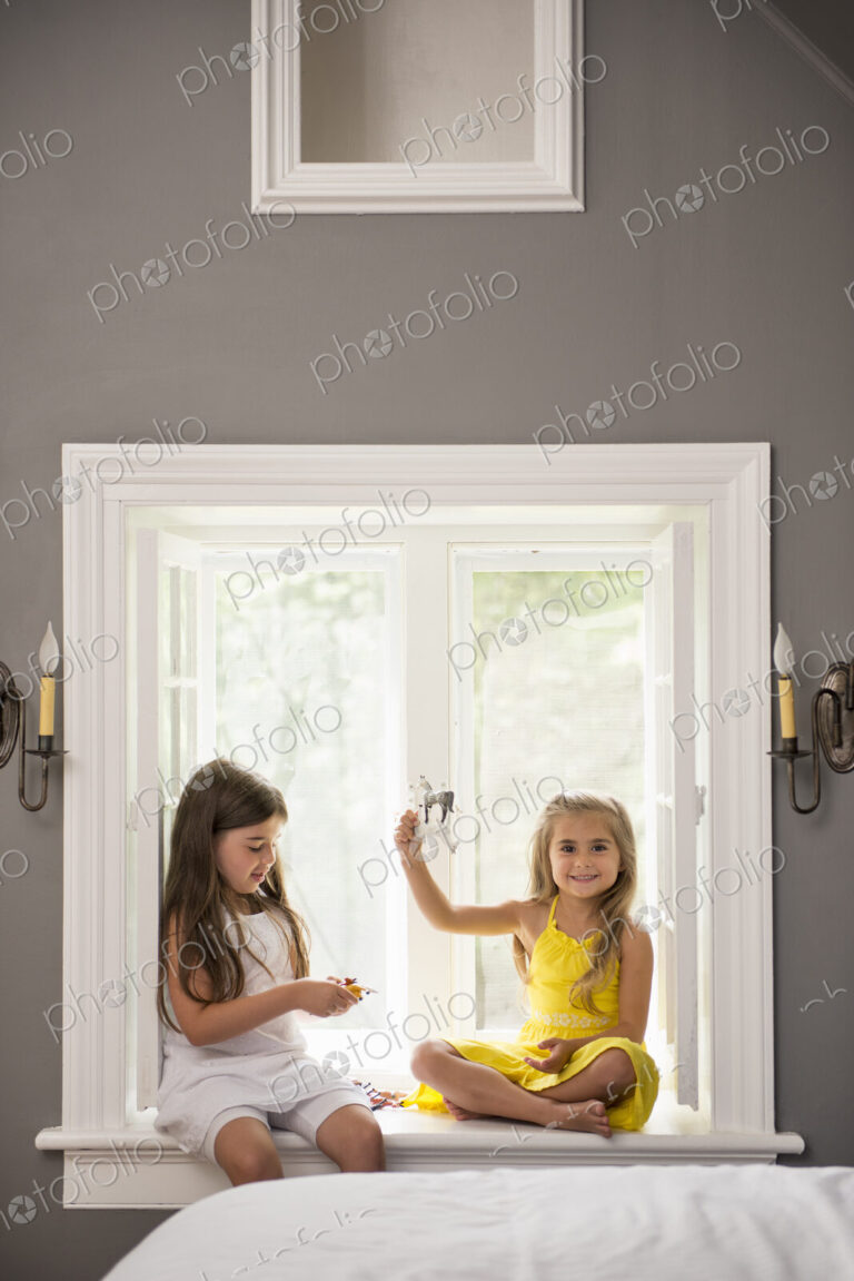 Two girls playing together, sitting on a window seat indoors.