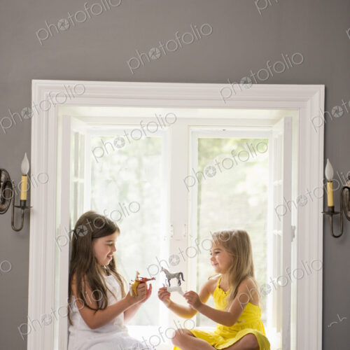 Two girls playing together, sitting on a window seat indoors.