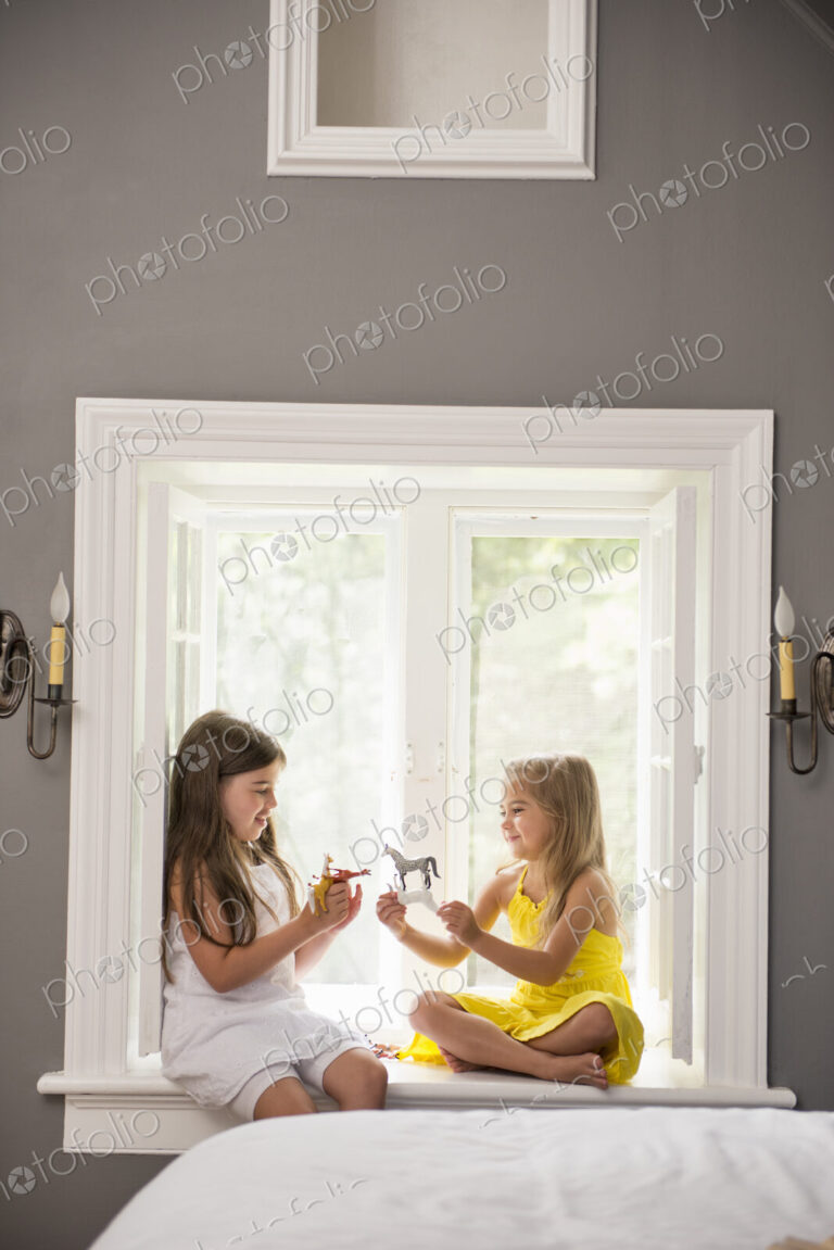 Two girls playing together, sitting on a window seat indoors.