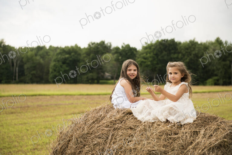 Two girls sitting on top of a haystack playing.
