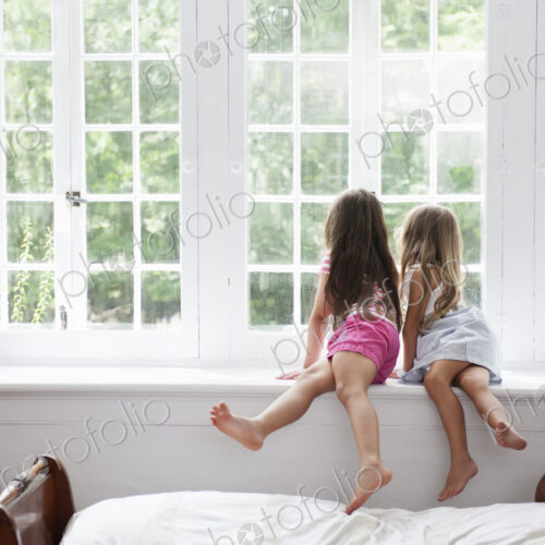 Two girls sitting side by side by a large window.