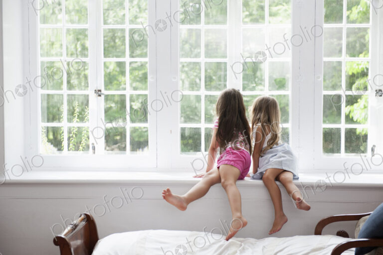 Two girls sitting side by side by a large window.