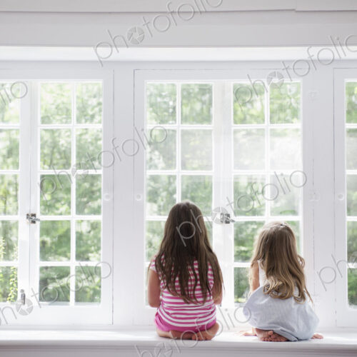 Two girls sitting side by side by a large window.